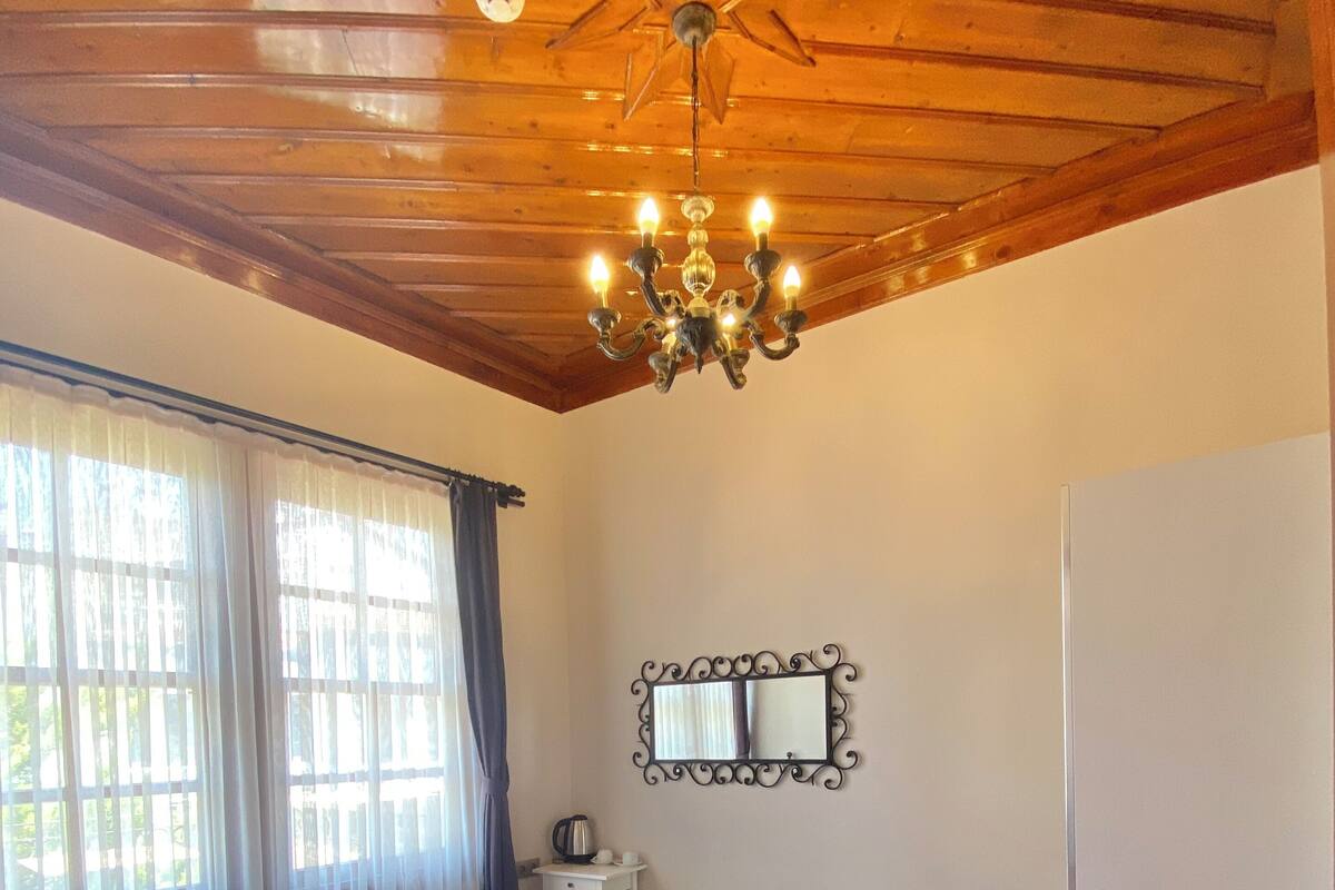 A bedroom with a wooden ceiling, a chandelier, a bed with white and brown bedding, a nightstand, a mirror, and a door.