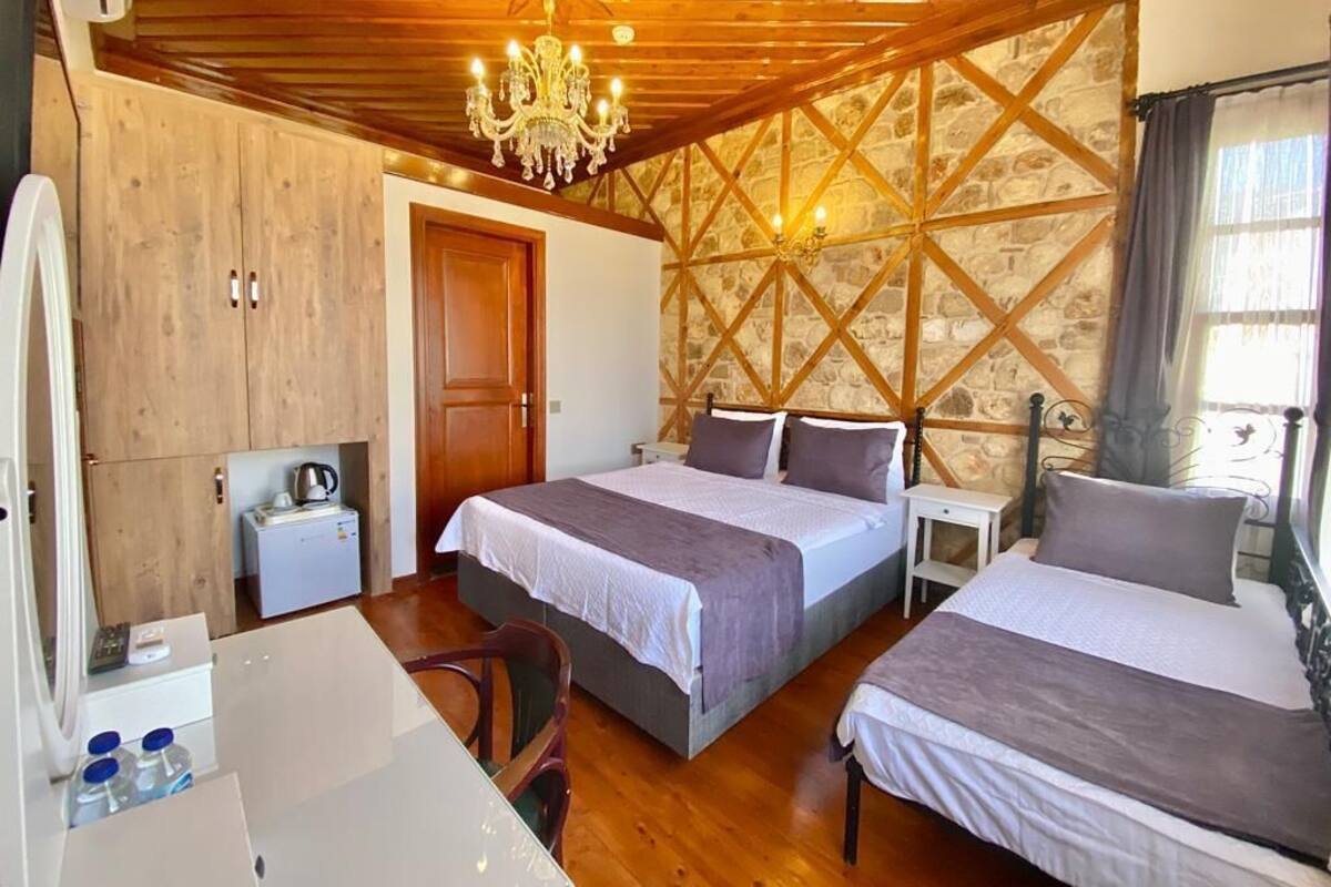 A room with two beds, a wooden ceiling, a stone wall, a chandelier, a desk, and a small kitchenette.