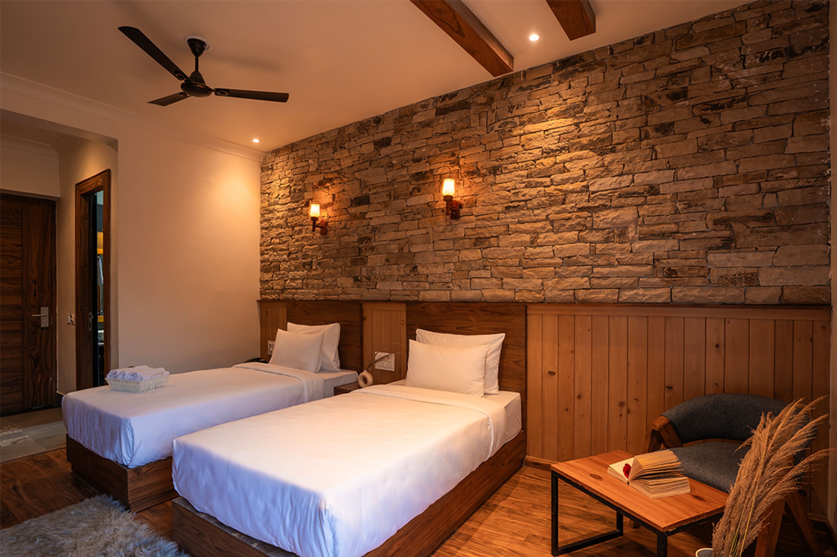 A room with two beds, a wooden ceiling fan, stone wall, and a small table with a book.