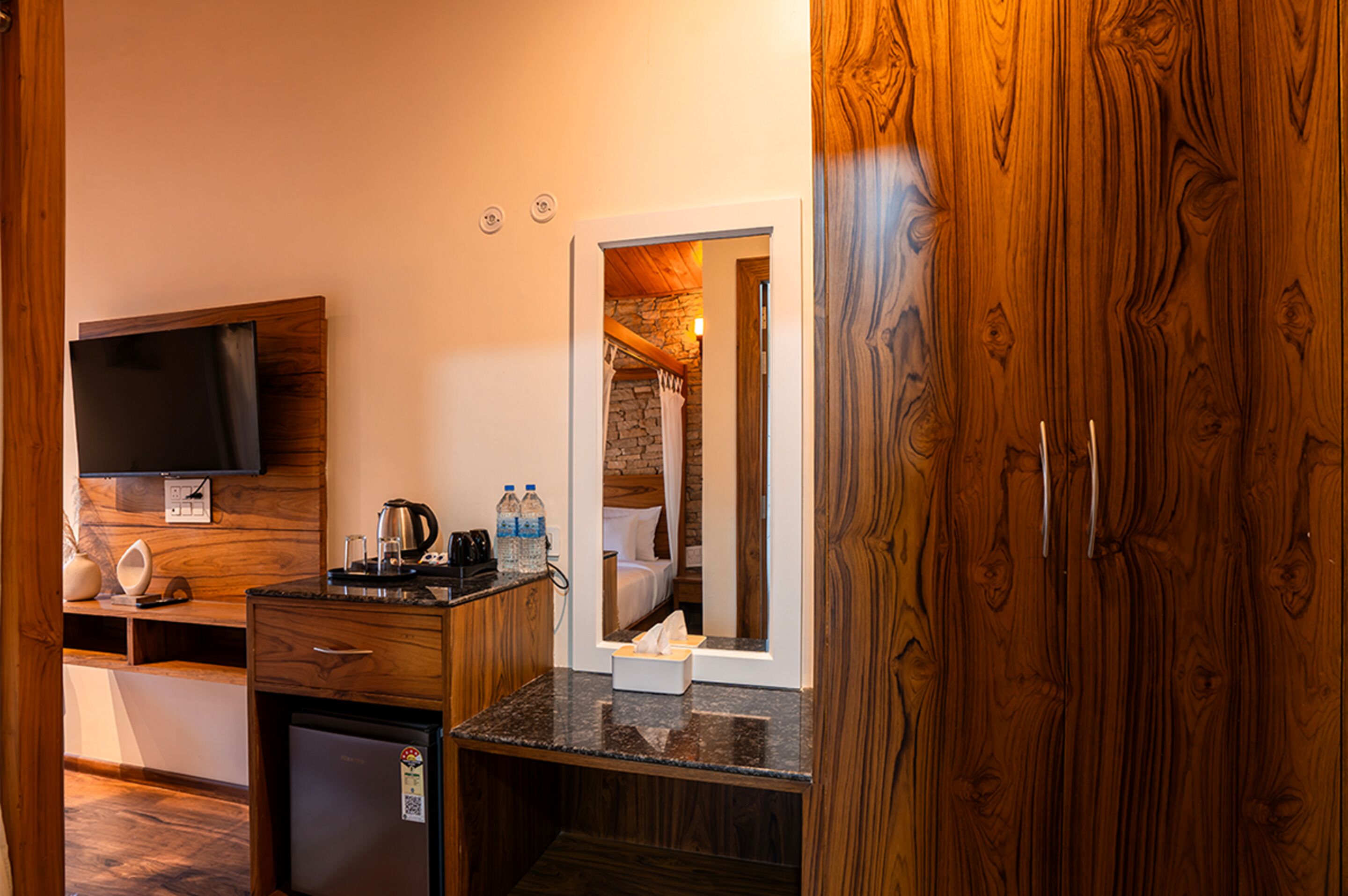 A compact hotel room with a kitchenette, a flat-screen TV, a small refrigerator, and a wooden wardrobe.