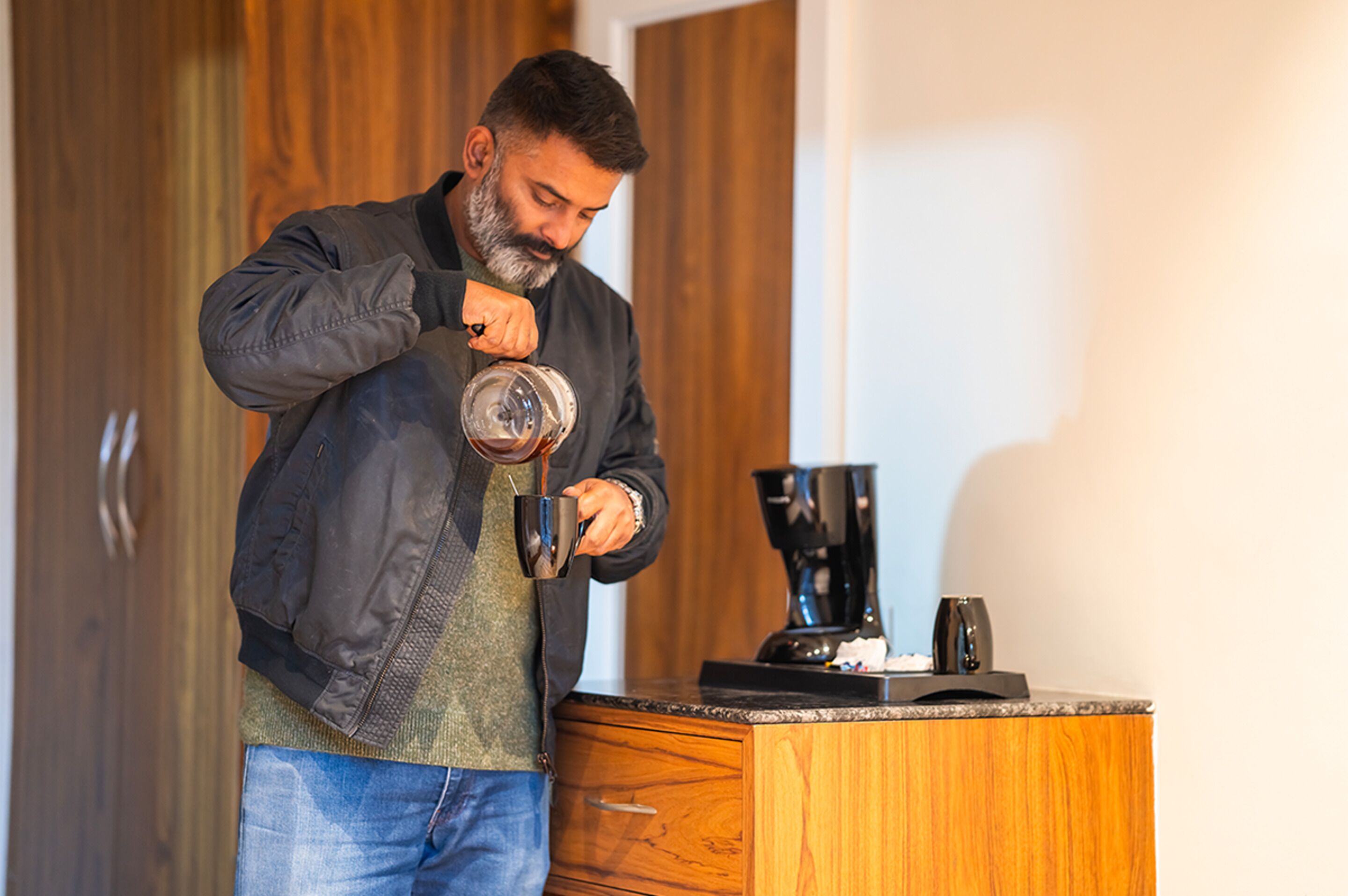 A person pouring coffee into a mug in a kitchen with wooden cabinets and a coffee maker on the counter.