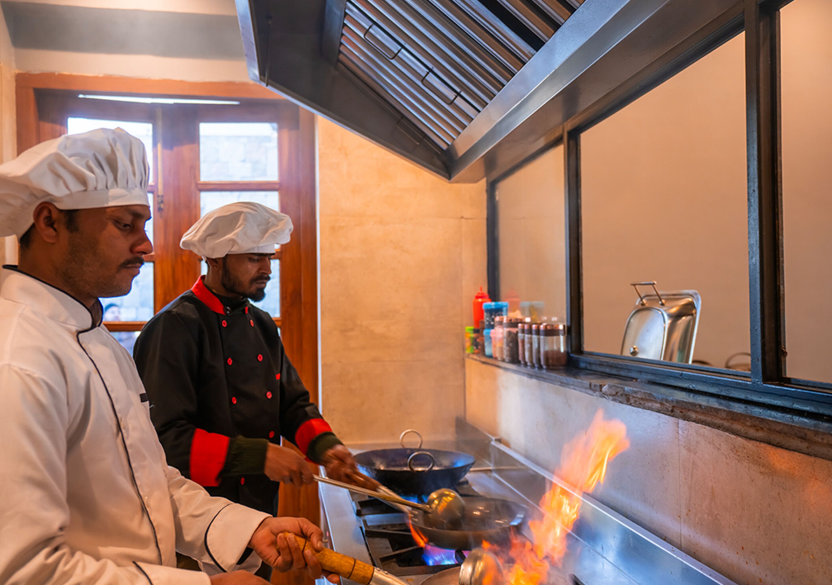 Two chefs cooking with a visible flame on a stove in a kitchen with wooden accents and a window.
