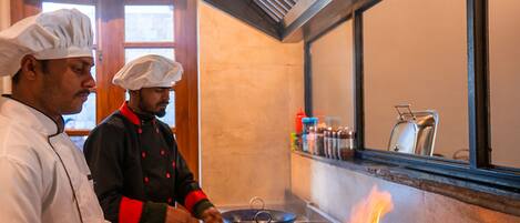 Deux chefs cuisinent sur une cuisinière, utilisant une flamme visible, dans une cuisine au décor chaleureux, avec des éléments en bois et une fenêtre.