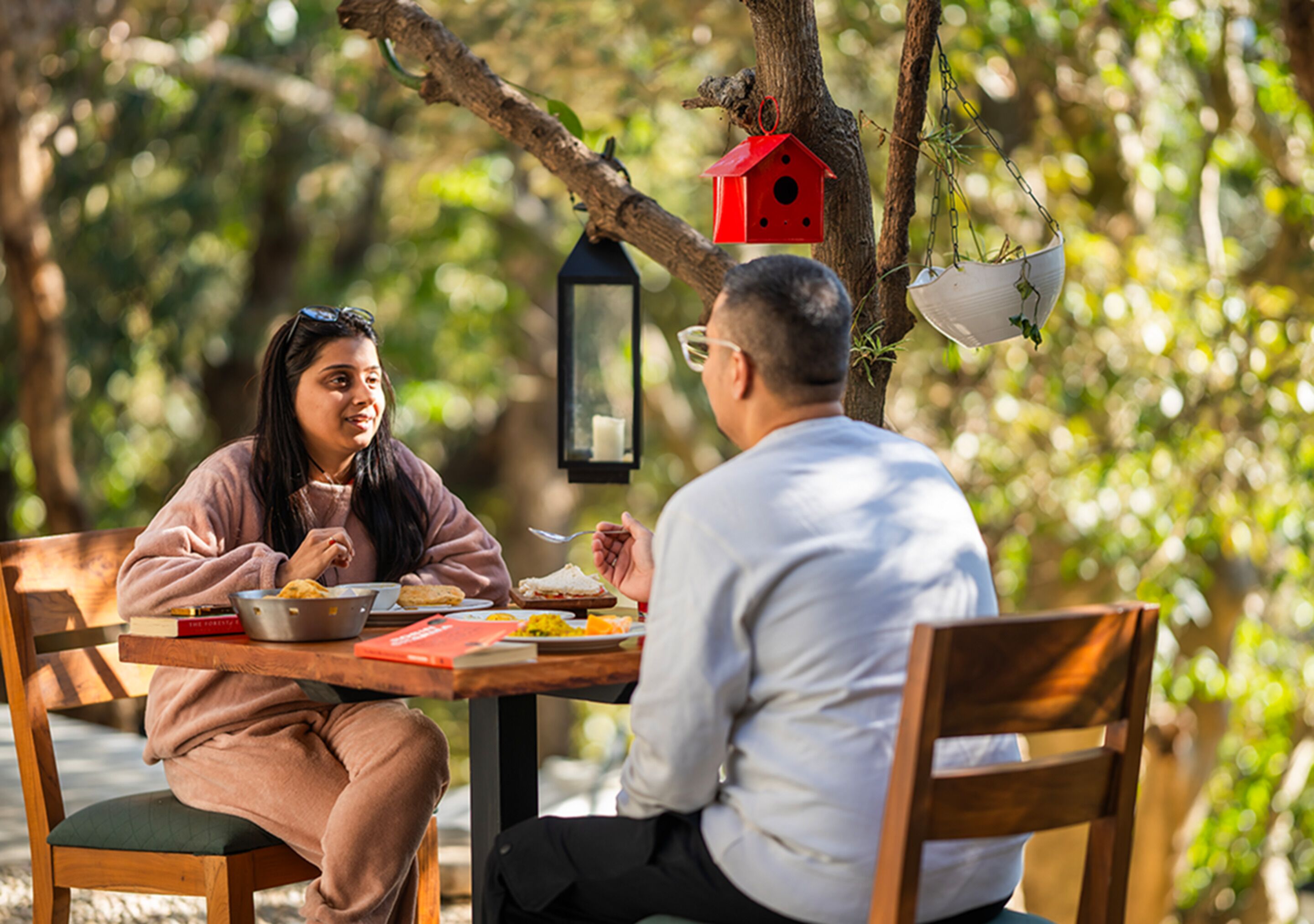 Two people sitting at an outdoor table with food, surrounded by trees and a birdhouse.