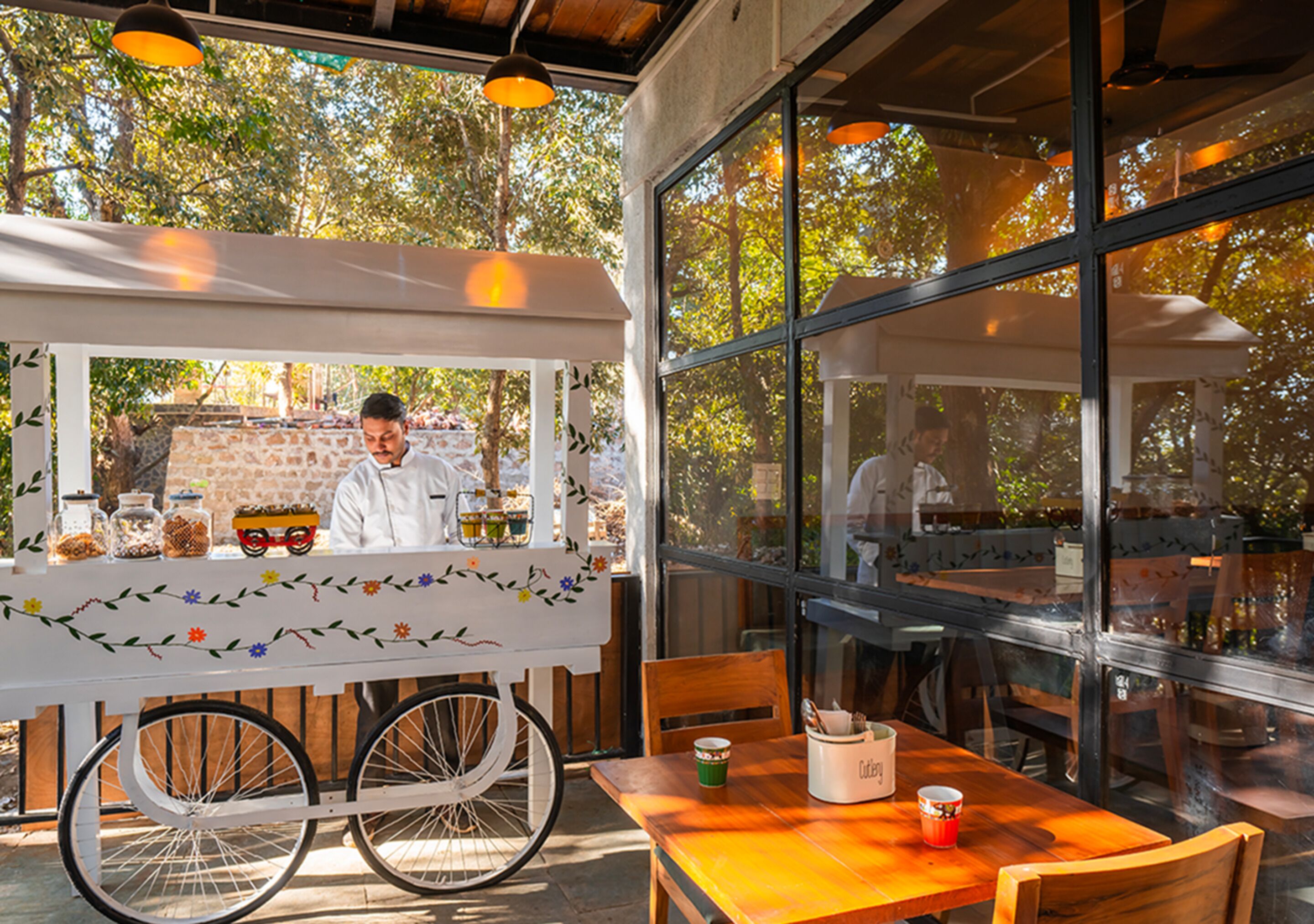 A food cart with a white canopy and a bicycle wheel base, positioned in front of a large window with a view of trees.