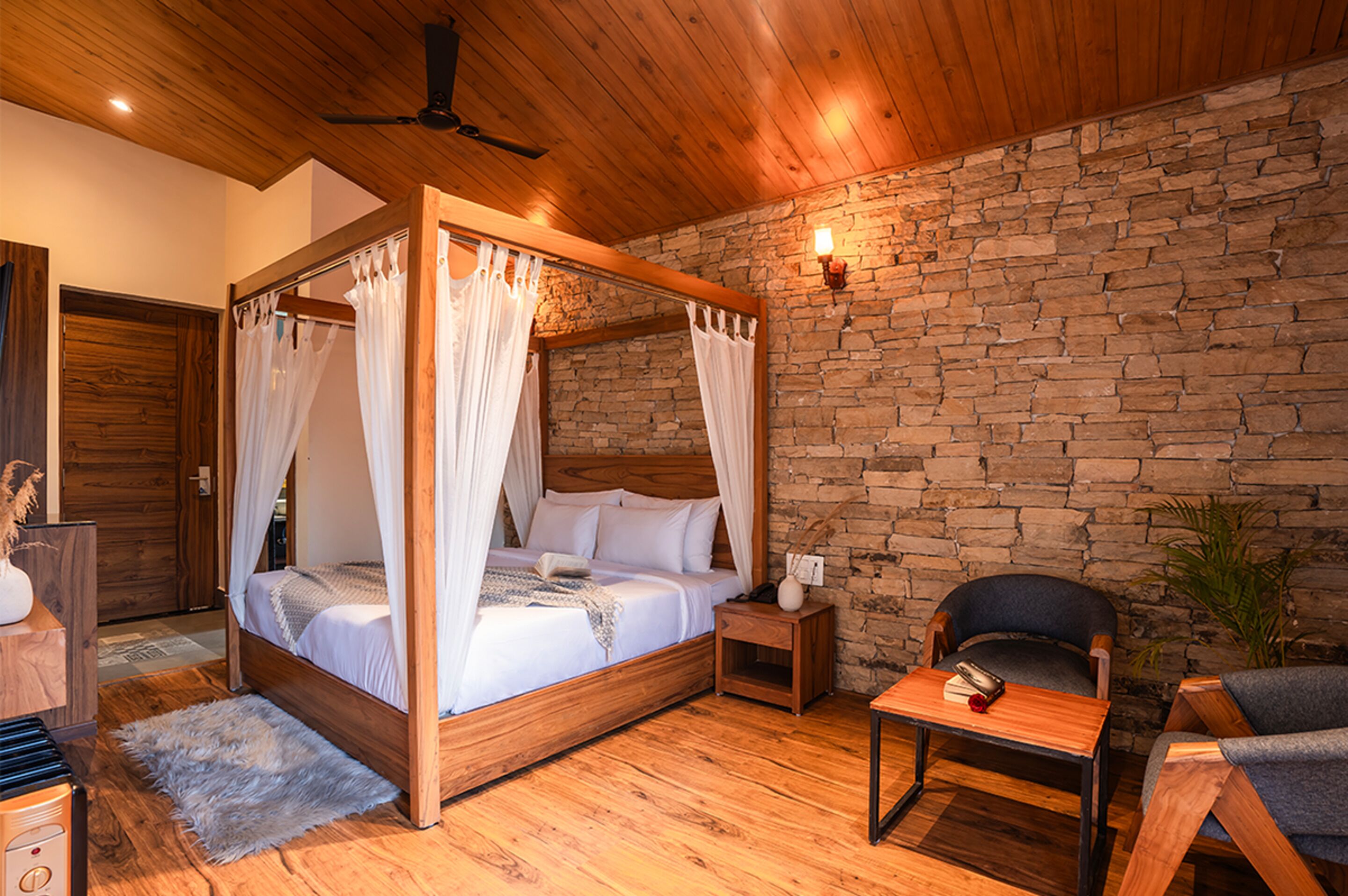 A four-poster bed with white bedding, a wooden nightstand, a chair, and a stone wall.