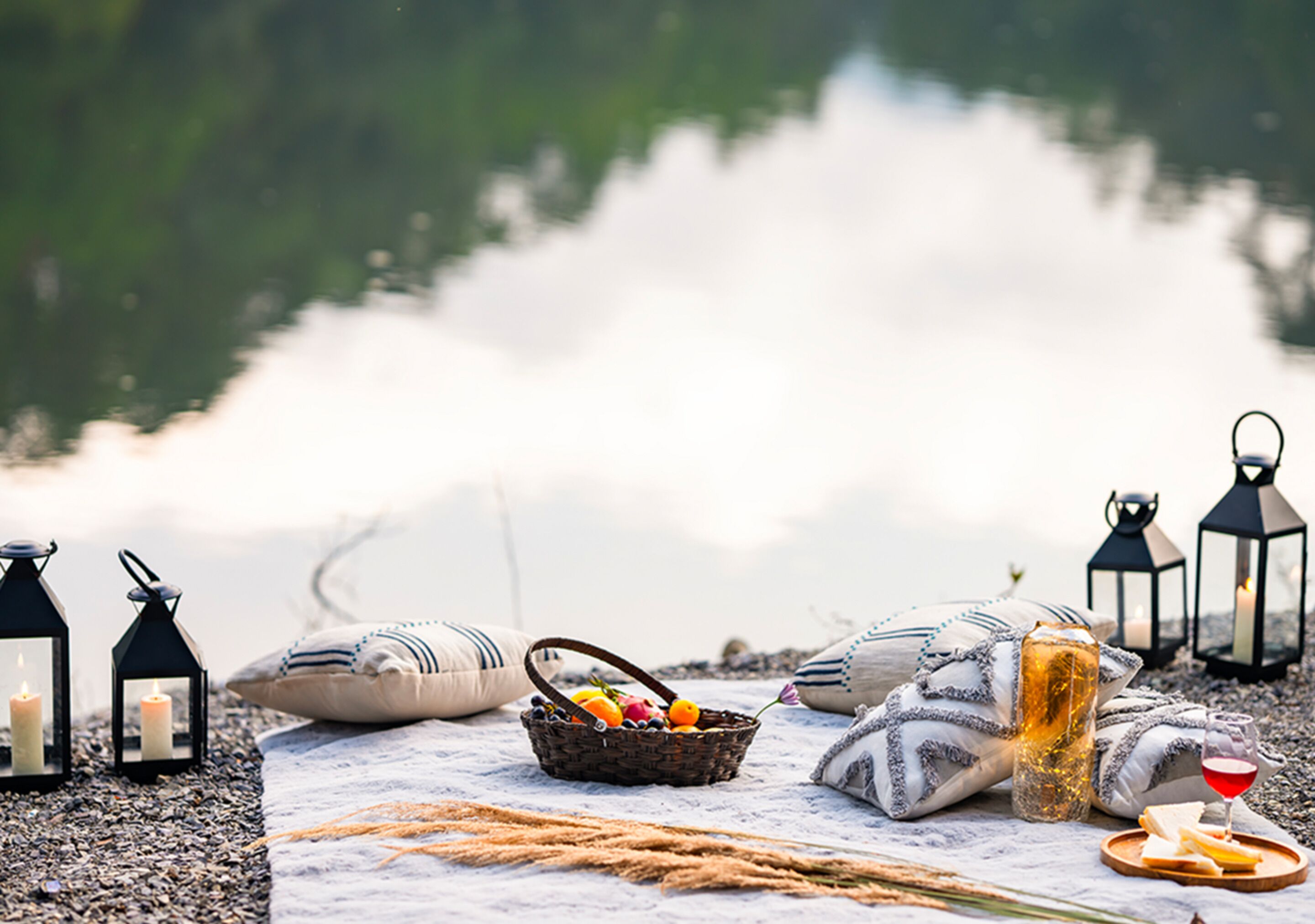 Un montaje para picnic junto a un lago, con faroles, almohadones, una cesta de fruta y una copa de vino.