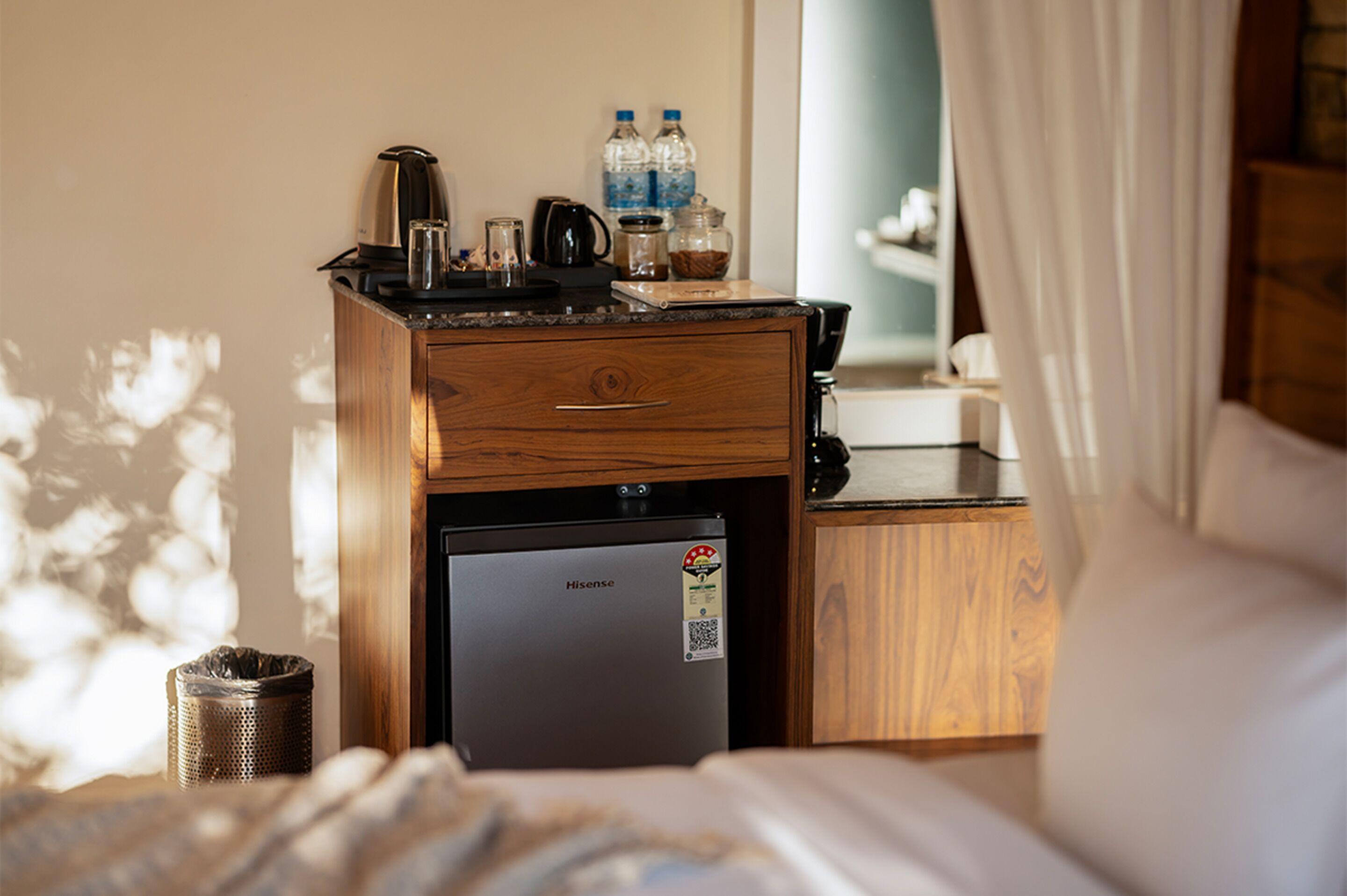 A compact hotel room with a wooden desk, a small refrigerator, a coffee maker, and a bed with a white comforter.