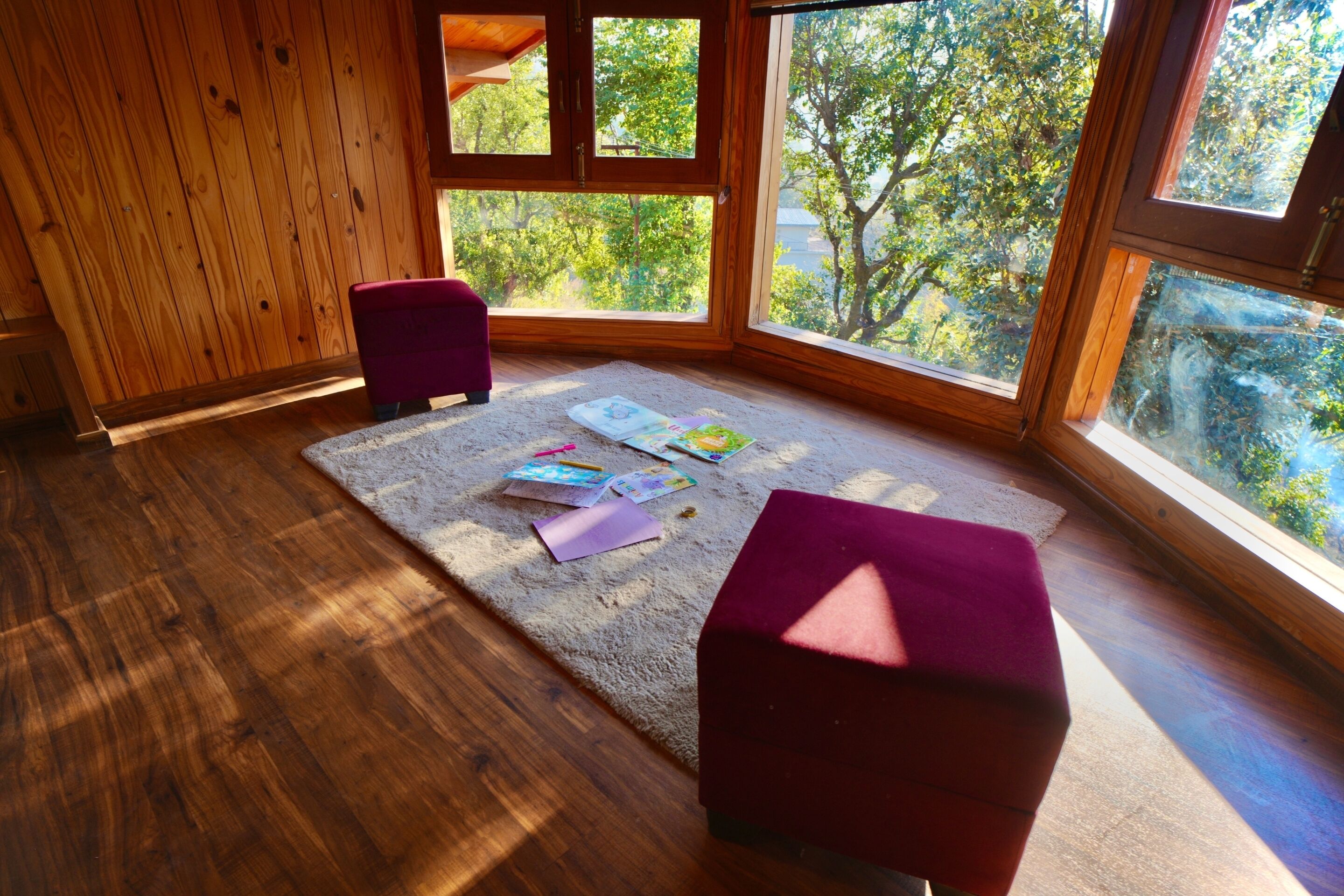 A room with wooden flooring, a large window with a view of trees, and two purple ottomans.