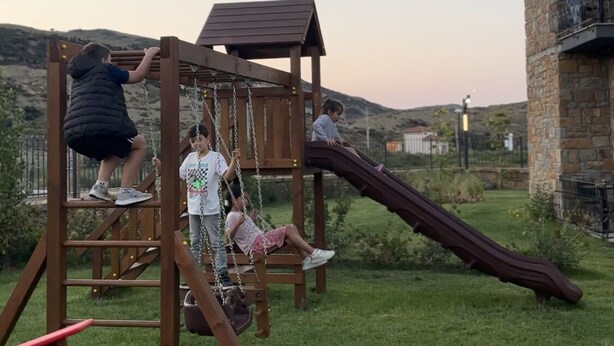 A playground with a slide and climbing structures, surrounded by a grassy area and a stone building. - Agios Hotel (Gökçeada)
