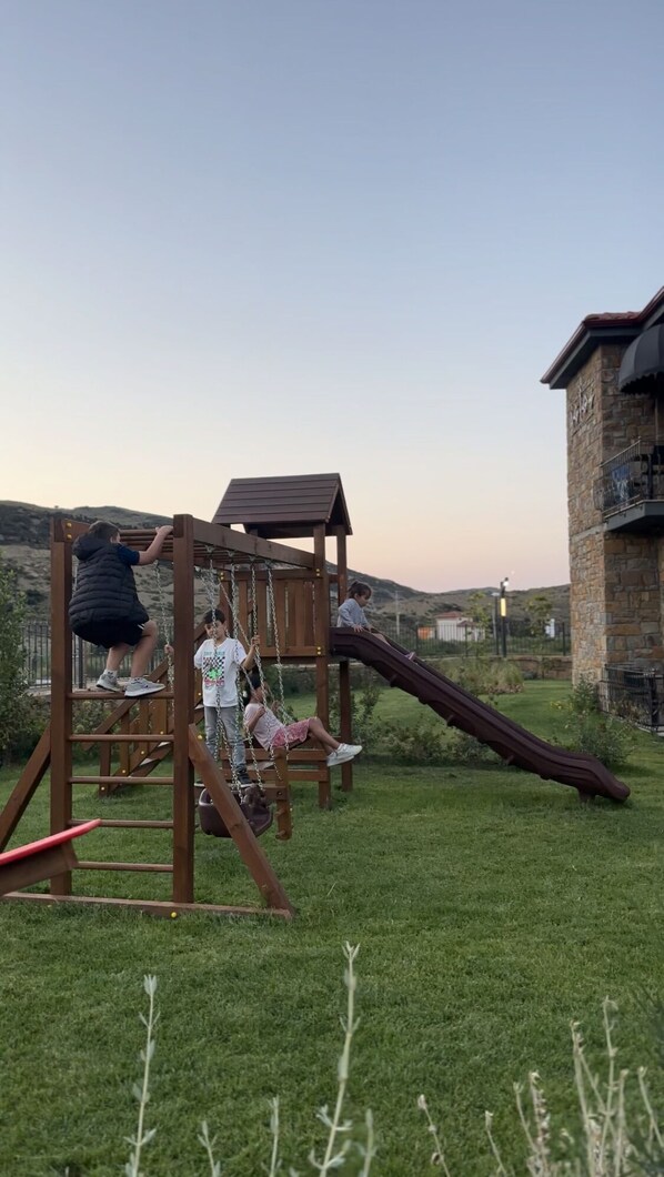 Un terrain de jeux avec un toboggan et des structures d’escalade, entouré d’une pelouse et bordé d’un bâtiment en pierre.