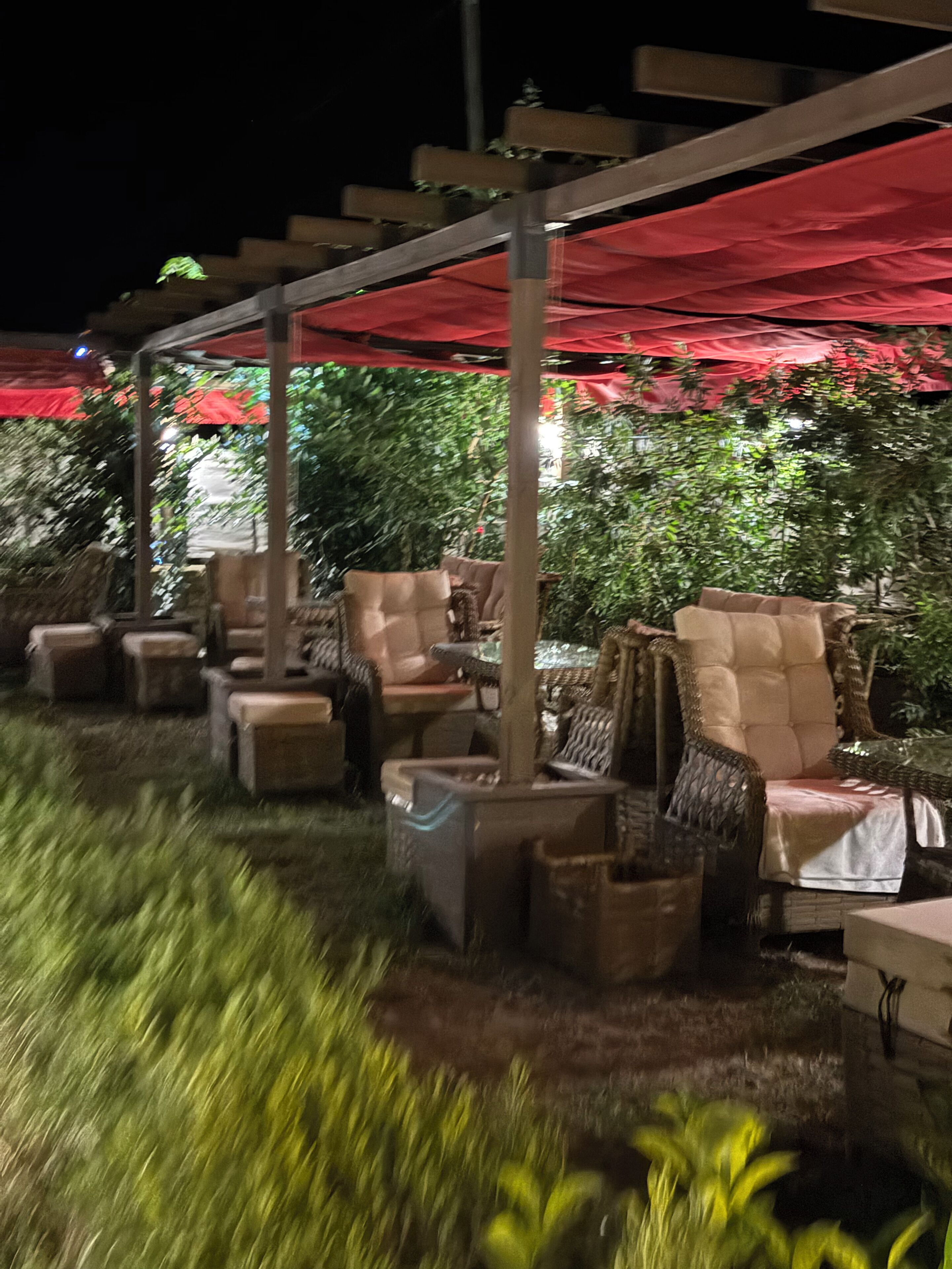 An outdoor seating area with multiple chairs and tables under a red canopy, surrounded by greenery.