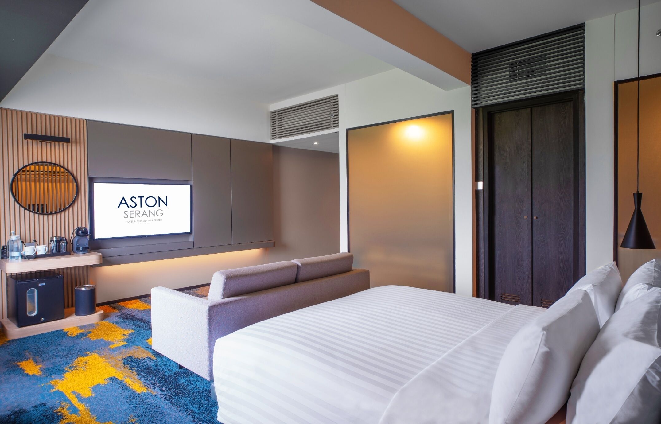 A modern hotel room with a large bed, a TV screen displaying 'ASTON SERANG', a coffee machine, and a minibar.