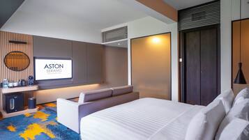 A modern hotel room with a large bed, a TV screen displaying 'ASTON SERANG', a coffee machine, and a minibar.