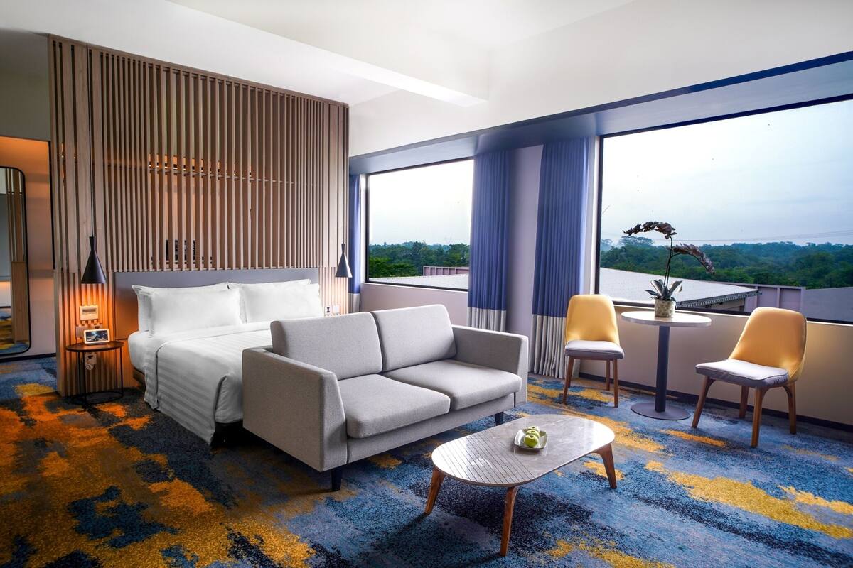 Ett modernt hotellrum med en stor säng, en soffa, två fåtöljer, ett litet bord och utsikt över grönområden genom stora fönster.