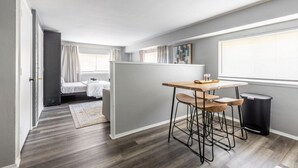 Ein kleines Zimmer mit Bett, einem Tisch mit Stühlen, einem Fenster mit Jalousien und einem Mülleimer.