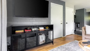 Ein modernes Wohnzimmer mit einem Flachbildfernseher an einer grauen Ziegelwand, einer schwarzen Unterhaltungsecke mit Büchern und Dekorationsobjekten, einer weißen Tür und einer durchgehenden Öffnung erkennbaren Küche.