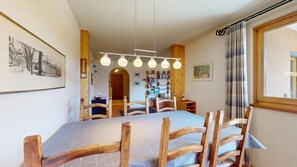 A dining area with a long table, wooden chairs, hanging pendant lights, a bookshelf, and a framed picture on the wall.