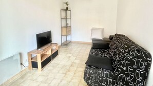 A living room with a TV, wooden entertainment unit, black and white patterned sofa, and a tall shelving unit. - Charming Studio for 2 in Ajaccio, Close to Airport and Amenities (Ajaccio)