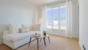 Una sala de estar moderna con un sofá blanco, una mesa redonda y vistas a un paisaje urbano a través de amplias ventanas.