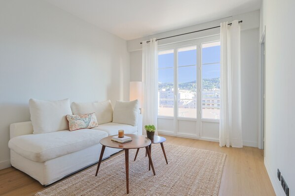 Een moderne woonkamer met een witte bank, een ronde salontafel en uitzicht op een stadsgezicht door grote ramen.
