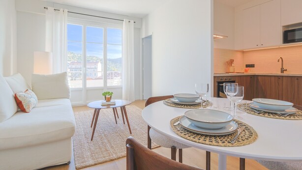 Una sala moderna con un sofá blanco, una mesa de comedor con platos y vasos, y una cocina con armarios de madera y un microondas.