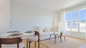 Una sala de estar moderna con una mesa de comedor preparada para cuatro personas, un sofá blanco y vistas de la ciudad a través de amplias ventanas.