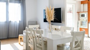 Un espace repas moderne avec une table et des chaises blanches, un vase contenant de l’herbe des pampas et un téléviseur à écran plat fixé au mur.