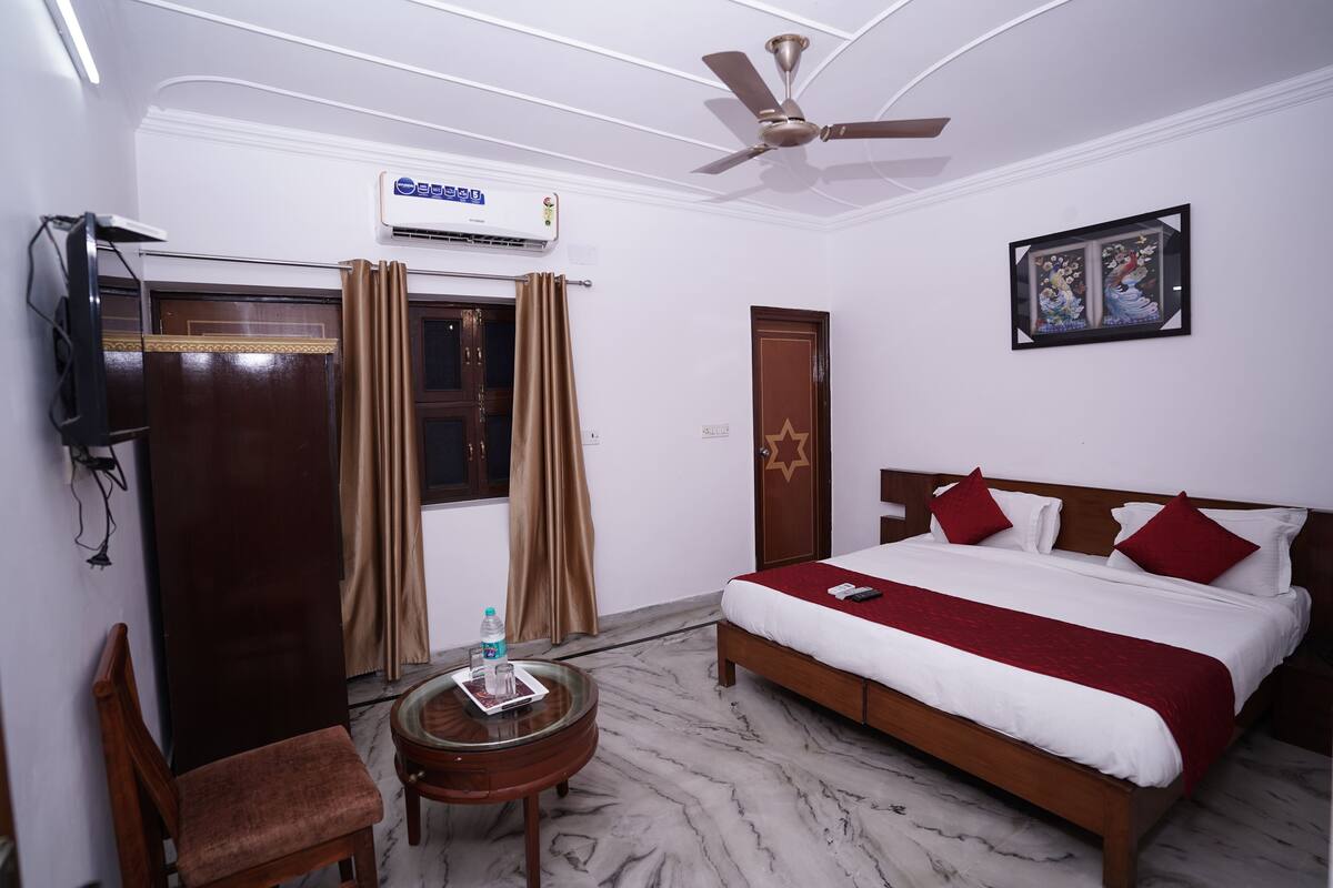 A hotel room with a bed, a ceiling fan, an air conditioner, a TV, a wooden wardrobe, a chair, a small table with a bottle, and a framed picture on the wall.