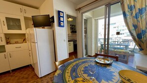 A compact kitchen with white cabinets, a white refrigerator, and a flat-screen TV on top. A dining table with a blue and yellow patterned tablecloth and a yellow plate is set with a wine glass.
