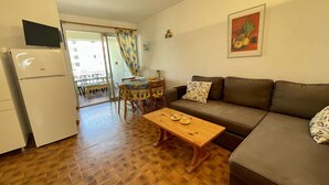 A compact living space with a sofa, a wooden coffee table, a dining table with chairs, a refrigerator, and a television.
