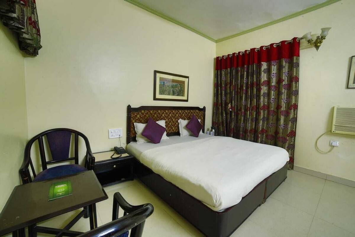 A hotel room with a bed, a desk, a chair, a nightstand, a telephone, and a wall-mounted air conditioning unit.