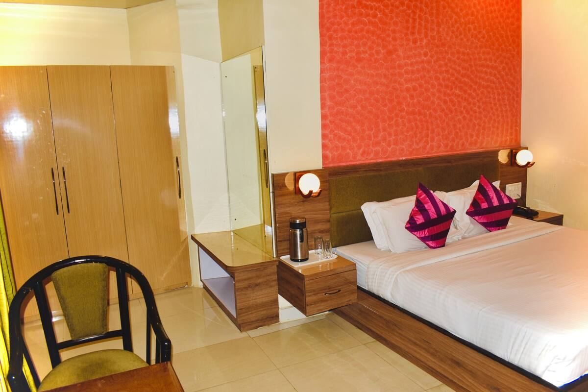 A hotel room with a wooden wardrobe, a bedside table with a lamp, a bed with white bedding and pink pillows, and a mirror above the dresser.