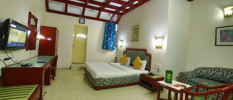 A hotel room with a bed, a desk, a chair, a sofa, a television, and a ceiling fan.