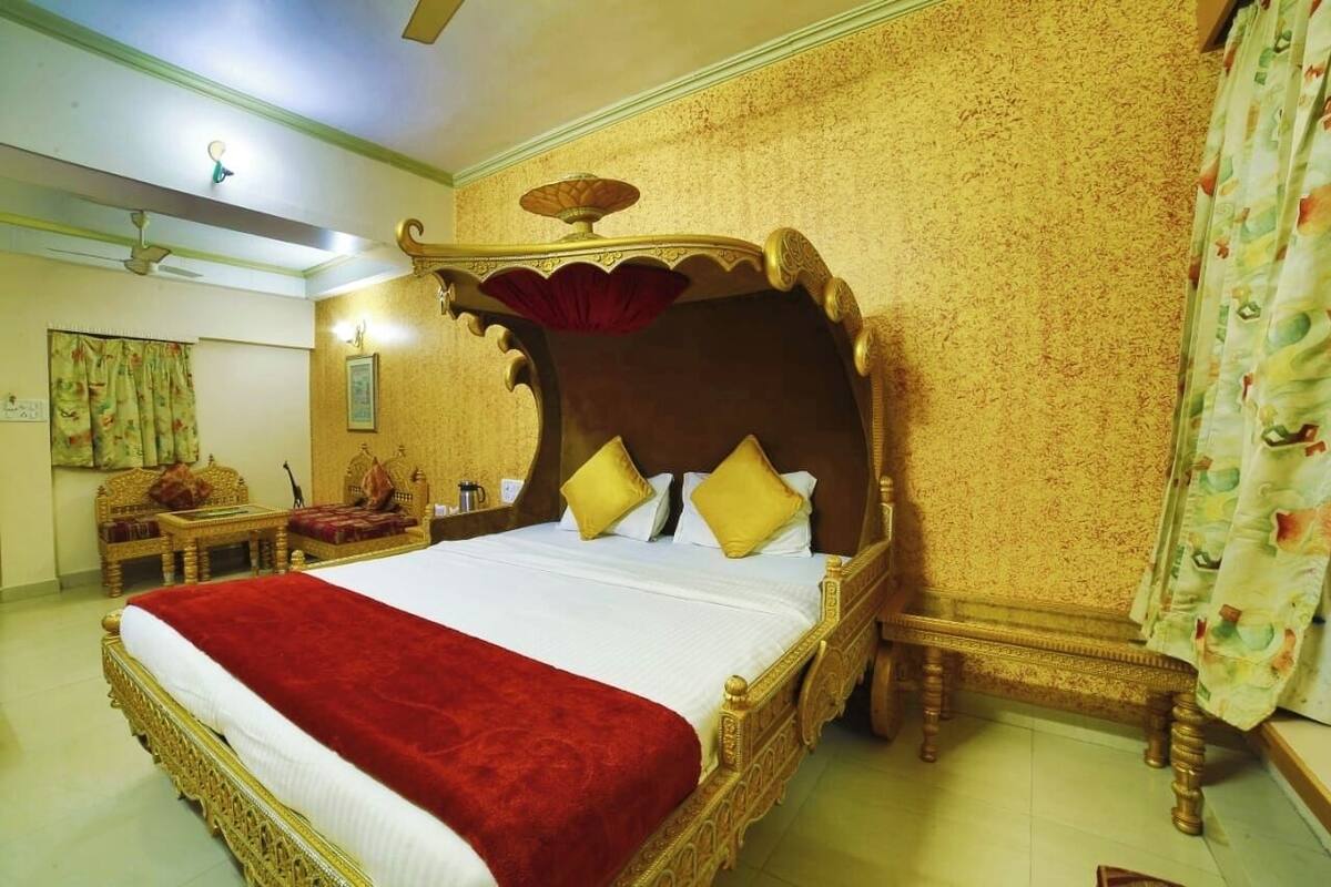 A large, ornate bed with a red bedspread and gold detailing, a wooden nightstand with a lamp, and a patterned curtain.