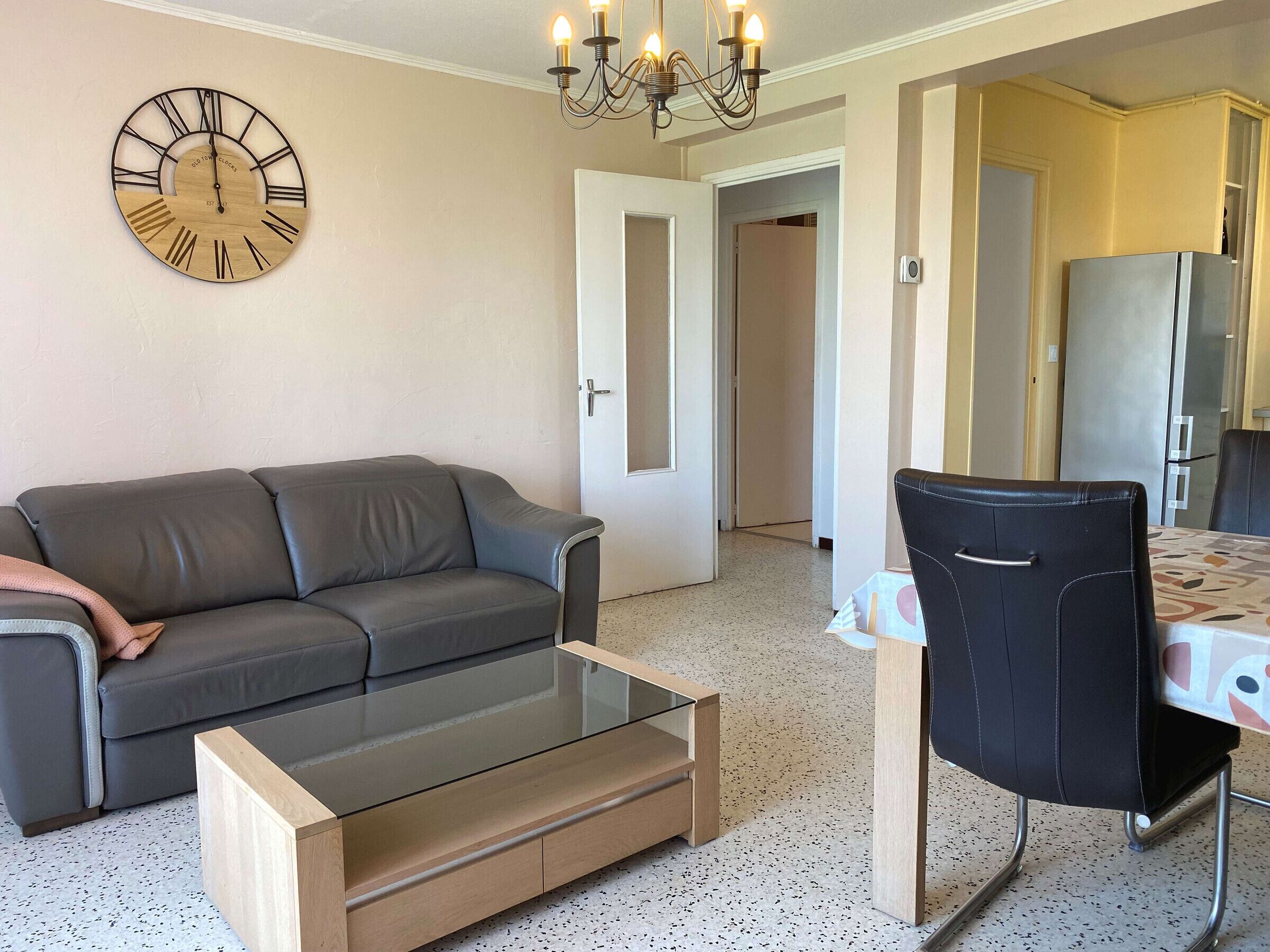 Un salon avec un canapé gris, une table basse en verre, un fauteuil noir, une horloge en bois et un lustre.