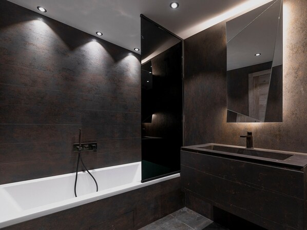 Un baño moderno con pared de azulejos oscuros, una bañera blanca, un espejo grande y luces empotradas en el techo.