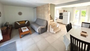 Ein Wohnzimmer mit einer Couch, einem Couchtisch und einem Essbereich mit Tisch und Stühlen. Im Hintergrund ist eine Küche mit Kühlschrank und Mikrowelle zu sehen.