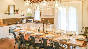 A dining area with a long table set for multiple people, kitchen cabinets, a microwave, and hanging pendant lights.
