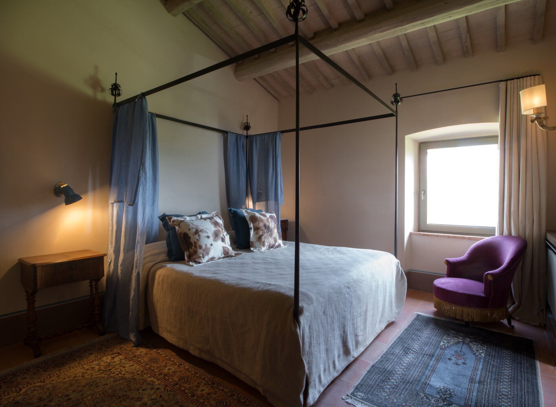 A four-poster bed with a white bedspread and brown and white patterned pillows, a wooden nightstand, a purple armchair, and a large window with curtains.