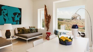 A modern living room with a sofa, a coffee table with a fruit bowl, a red stool, and a large window with a view of a landscape. - Villa Calipso - Homelike Villas (Nardò)
