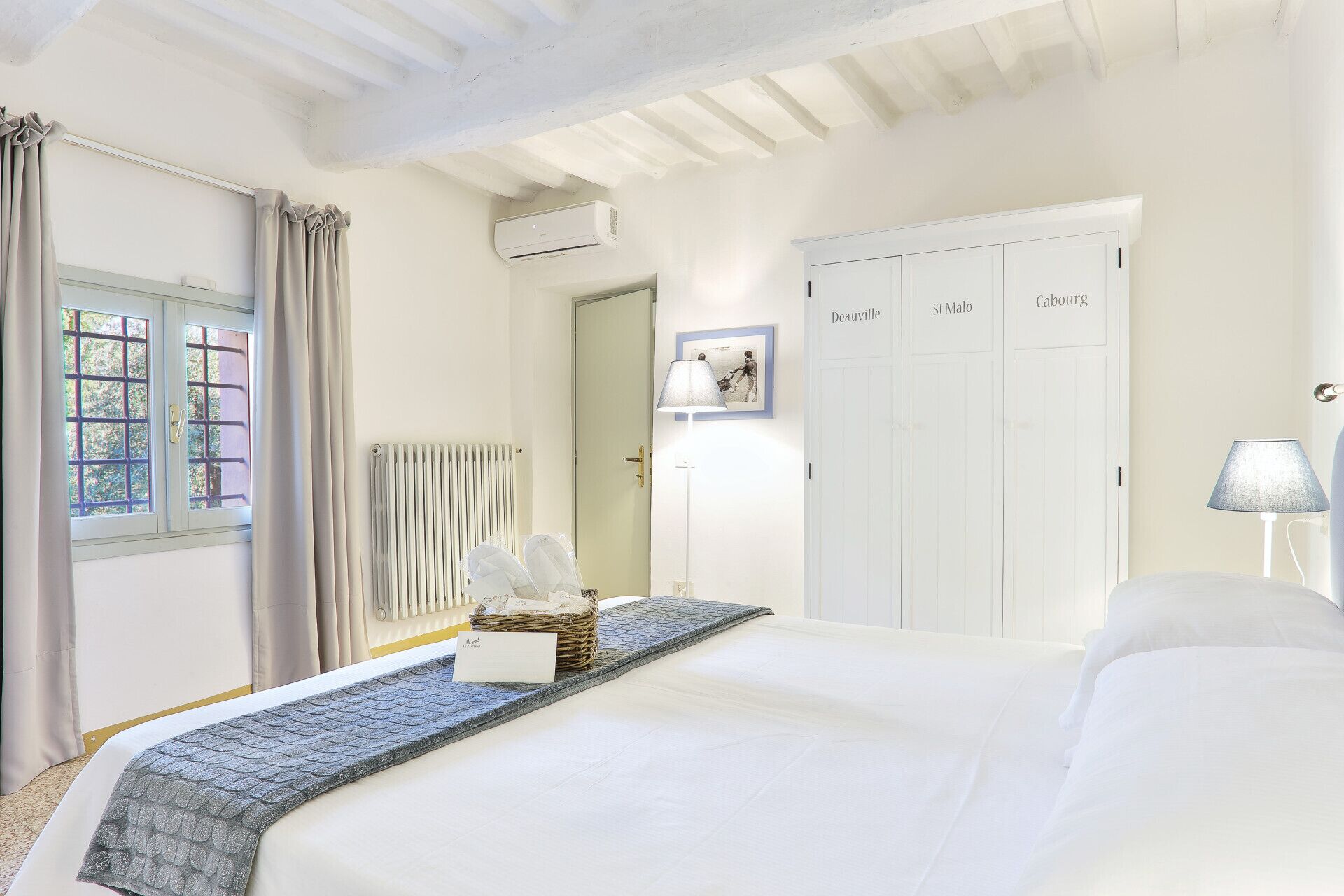 A hotel room with a bed, a radiator, a wardrobe with doors labeled 'Deauville', 'St Malo', and 'Cabourg', and a lamp on the right side.
