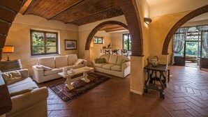 Amplia sala de estar con dos sofás, una mesa de centro y una chimenea. La habitación presenta techos de ladrillo visto y ventanas grandes con cortinas.