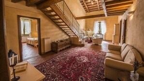 Un salón espacioso con techo alto, vigas de madera a la vista, un tapete grande de diseño rojo, un sofá y una escalera que lleva a otra habitación.