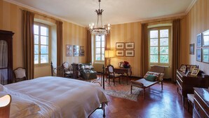 Une pièce bien éclairée, avec un grand lit, un bureau doté d’une lampe et une chaise. Plusieurs fenêtres, équipées de rideaux, donnent sur l’extérieur, et un lustre est suspendu au plafond.