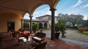Une terrasse extérieure avec des meubles en osier et des plantes en pot, un bâtiment avec un toit en tuiles en arrière-plan, et un chemin de gravier menant à ce bâtiment.