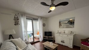 A living room with a sofa, a coffee table, a TV, a cabinet, and a window with curtains. - Cozy Studio for 2 Near Beach with Private Parking (Cap d'Agde)