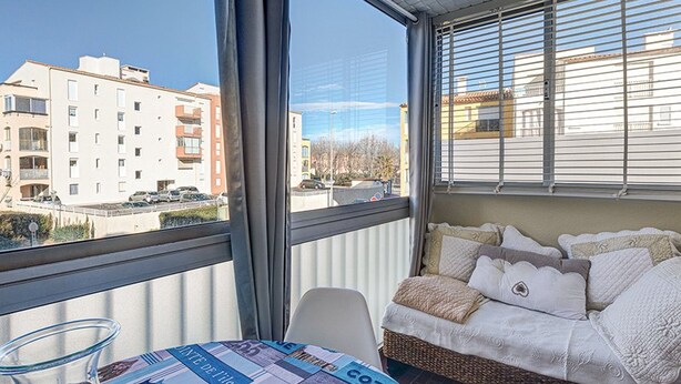 A balcony with a wicker sofa, a glass table with a candle, and a view of buildings. - Cozy Studio for 2 Near Beach with Private Parking (Cap d'Agde)