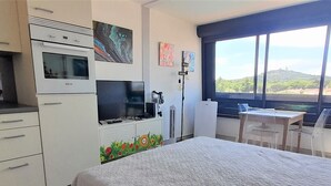 A bedroom with a bed, a TV, a desk, and a window with a view of greenery. - Cozy Studio for 2 with A/C, Pool Access, & Parking in Agde (Cap d'Agde naturisme)