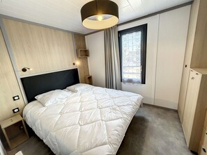 A modern hotel room with a large bed, a wardrobe, a nightstand, and a window with curtains. - Charming Chalet, 4 Sleeps, Heated Pool, Terrace, Close to Beach (Jullouville)